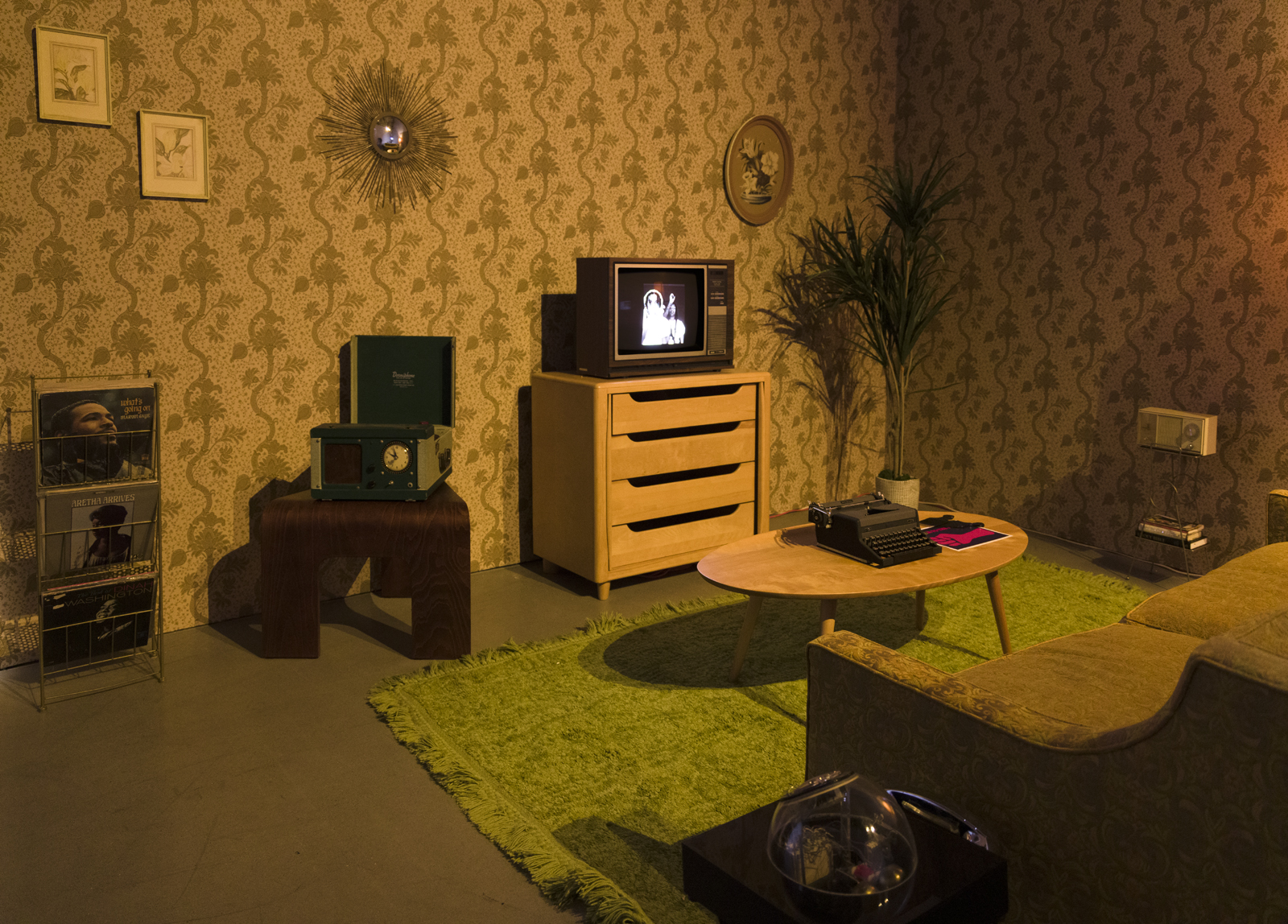 installation view of blitz bazawule's a moment in time at uta artist space, with dark, yellow-toned room with retro TV, couch, wallpaper and magazine rack