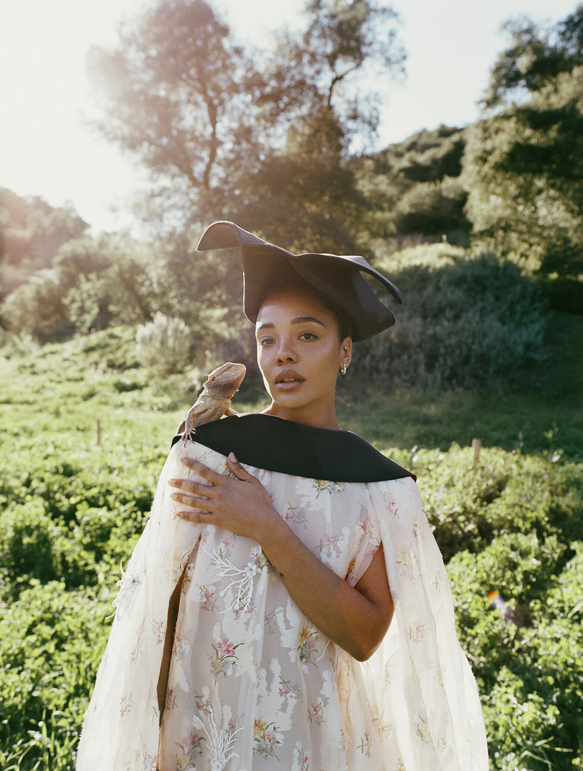 Actress Tessa Thompson poses in a grassy field wearing a leather hat, a floral dress, and carrying a bearded dragon on her shoulder.